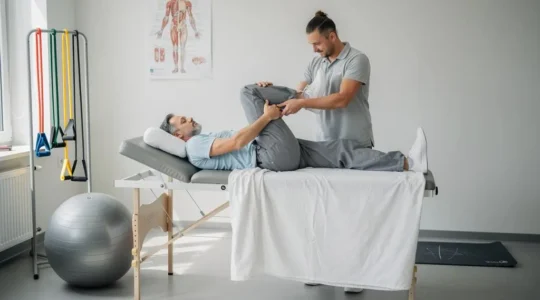 Un kinésithérapeute guide un patient lors d'un exercice de flexion du genou sur une table de rééducation dans un cabinet lumineux.